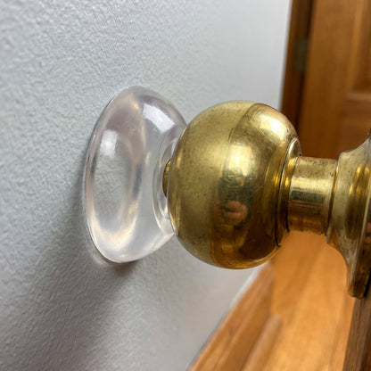 A close-up of a clear silicone wall protector adhered to a light gray wall, positioned behind a shiny brass doorknob.