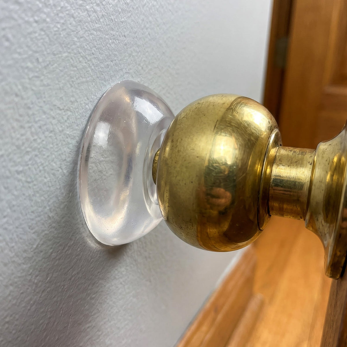 A close-up of a clear silicone wall protector adhered to a light gray wall, positioned behind a shiny brass doorknob.