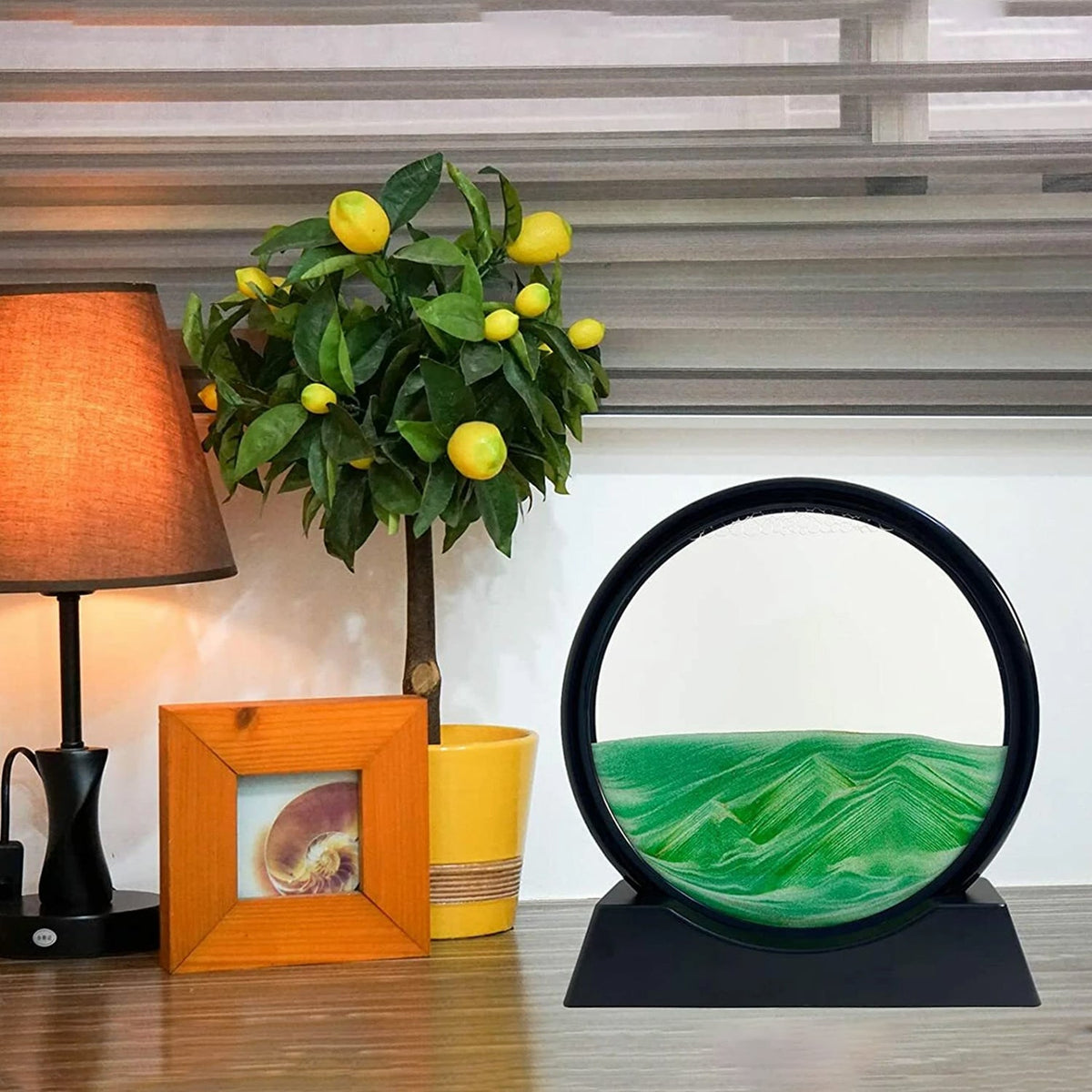A black-framed circular sand art display with green sand forming waves, next to a small lemon tree and a lamp.