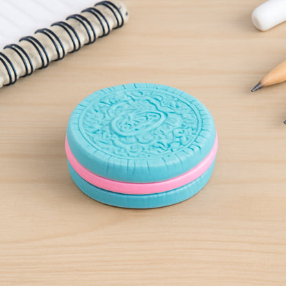 A blue and pink cookie-shaped eraser rests on a wooden desk next to a spiral notebook and a pencil.