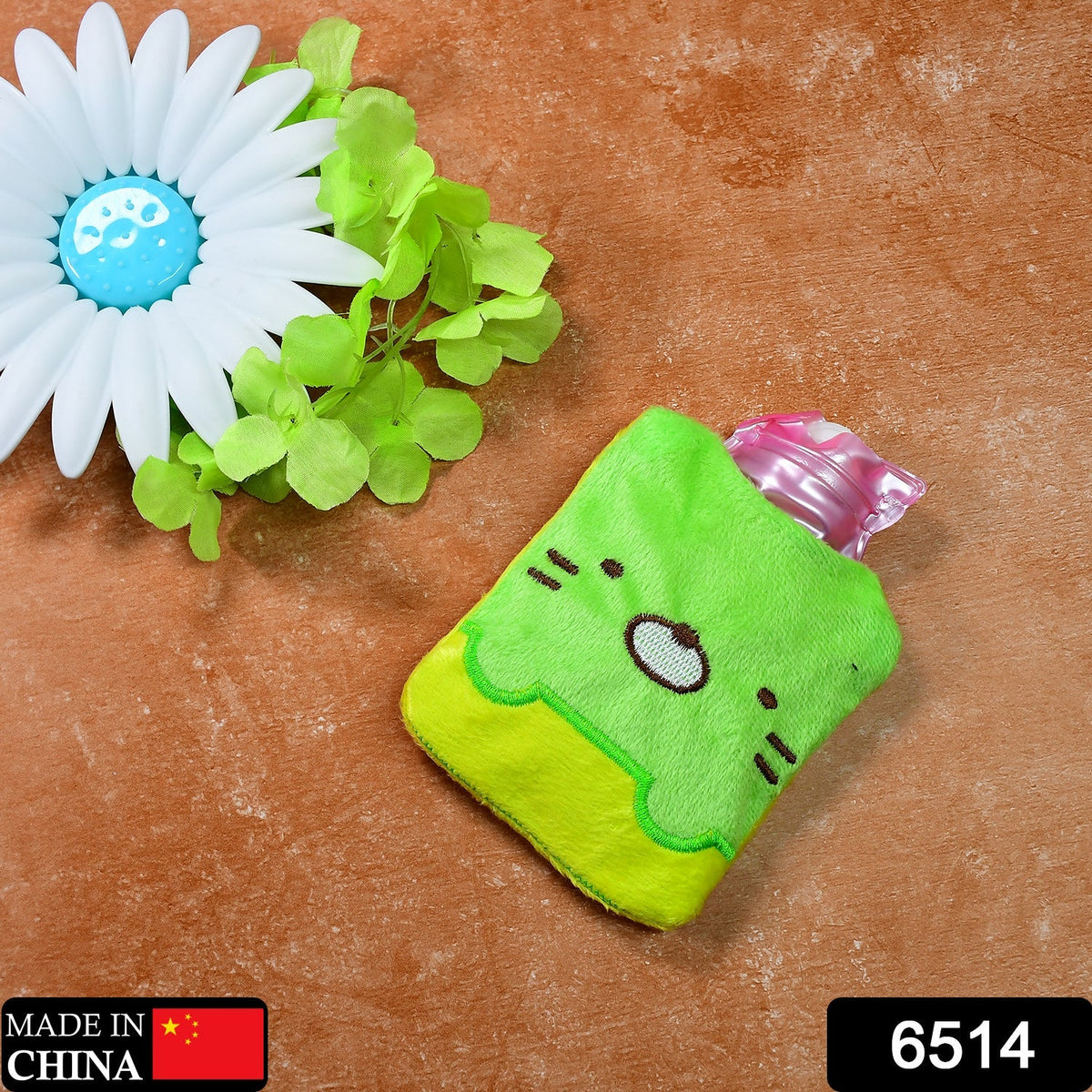 A green and yellow kitty hot water bag lies next to a white flower and green leaves on a brown surface.