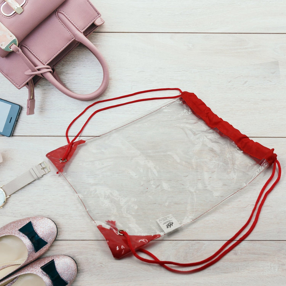 Clear drawstring bag with red cords and accents, surrounded by a pink handbag, sparkly shoes, and a watch on a wooden floor.