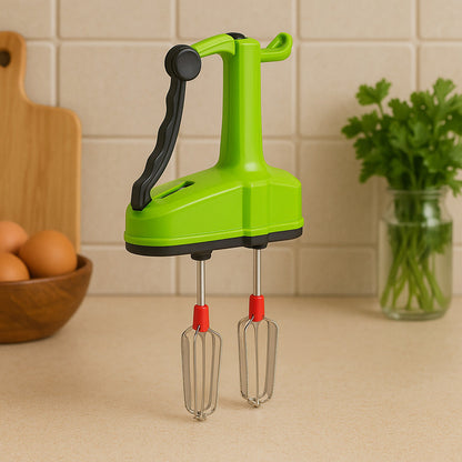 A green manual hand blender with black handle and red-tipped whisks sits on a kitchen counter with eggs and parsley in the