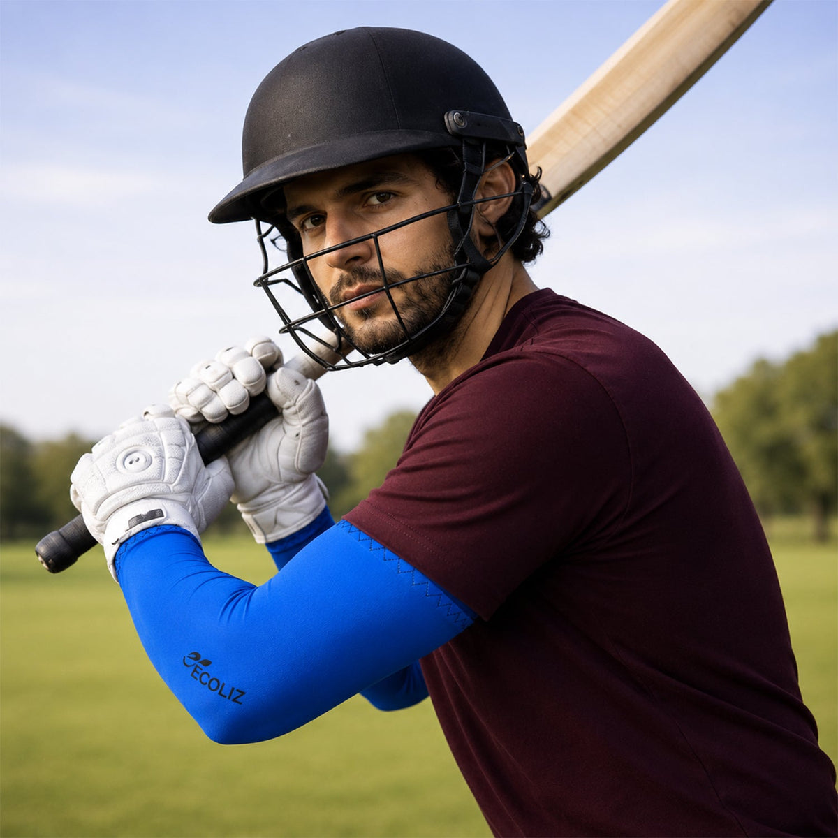 A man in a cricket helmet and white gloves holds a bat, wearing blue arm sleeves and a maroon shirt.