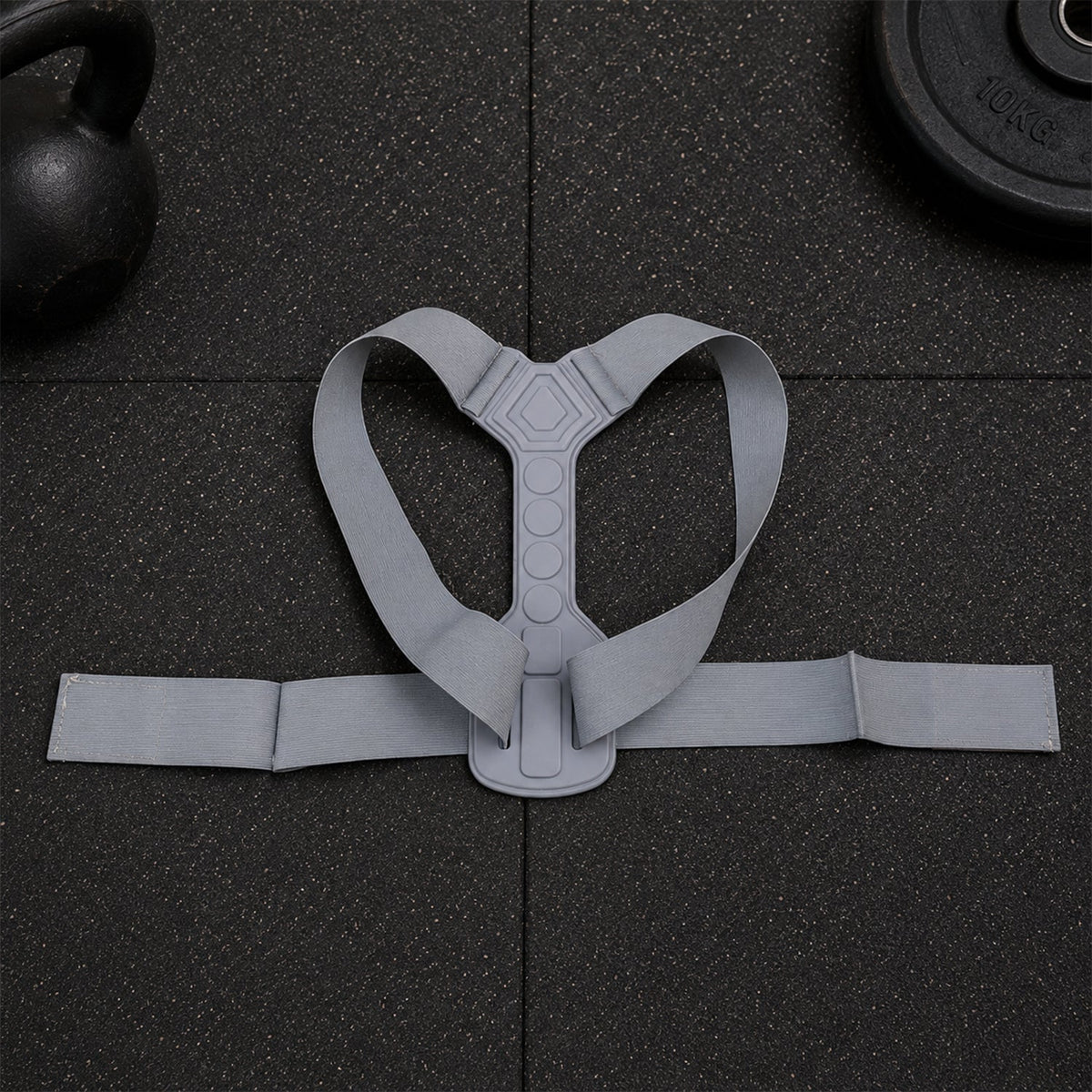 A grey posture corrector belt with a central spine support and shoulder straps laid flat on a dark textured surface.