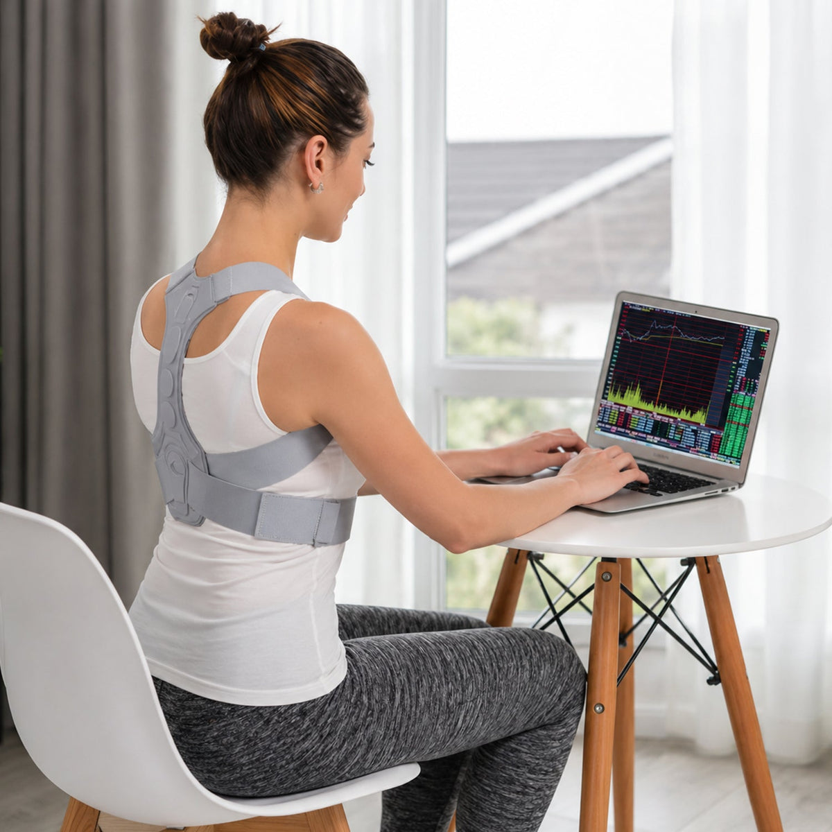 Woman wearing a grey posture corrector, typing on a laptop at a white table, with a window in the background.
