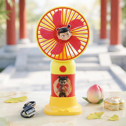 A red and yellow mini fan with a cartoon character and stand, next to a small peach, leaves, and a box.