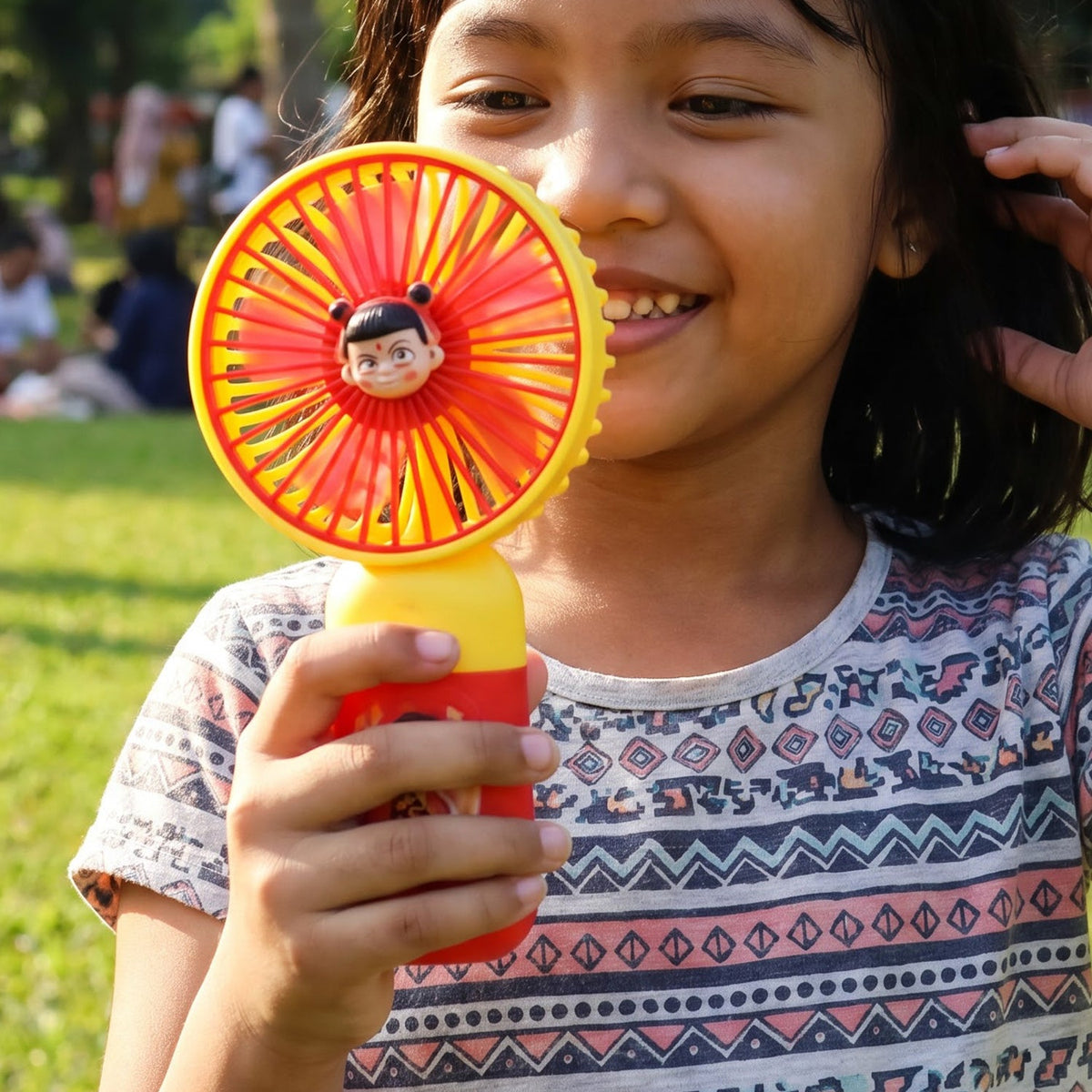 A young girl smiling and holding a red and yellow mini fan with a cartoon character face in the center. – view 7