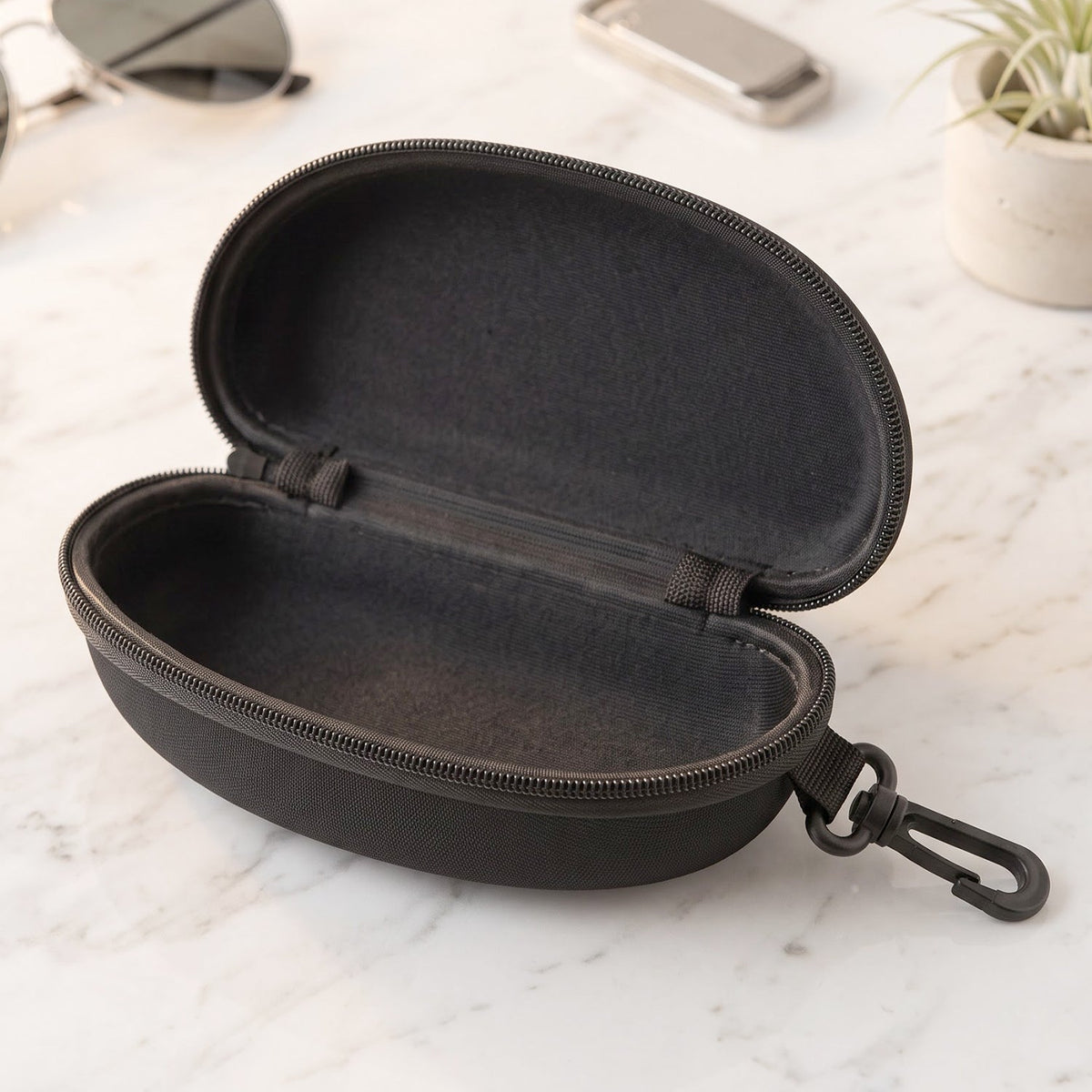 Empty open black hard shell sunglasses case with a zipper and clip, on a white marble surface.