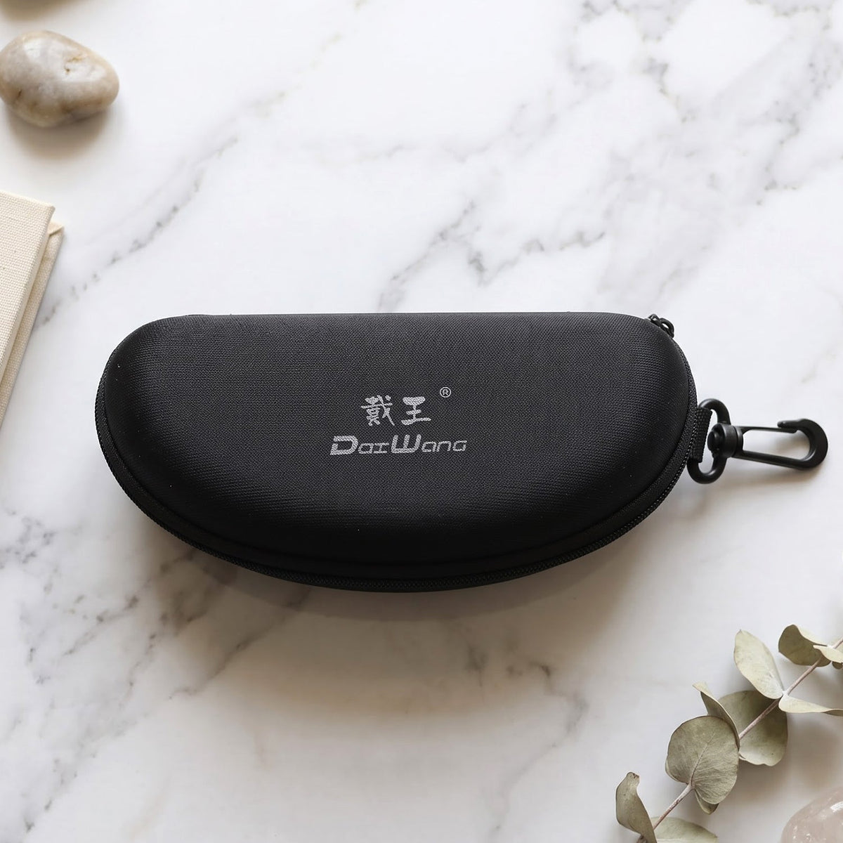 Closed black hard shell sunglasses case with a white logo and clip, resting on a white marble surface with foliage.