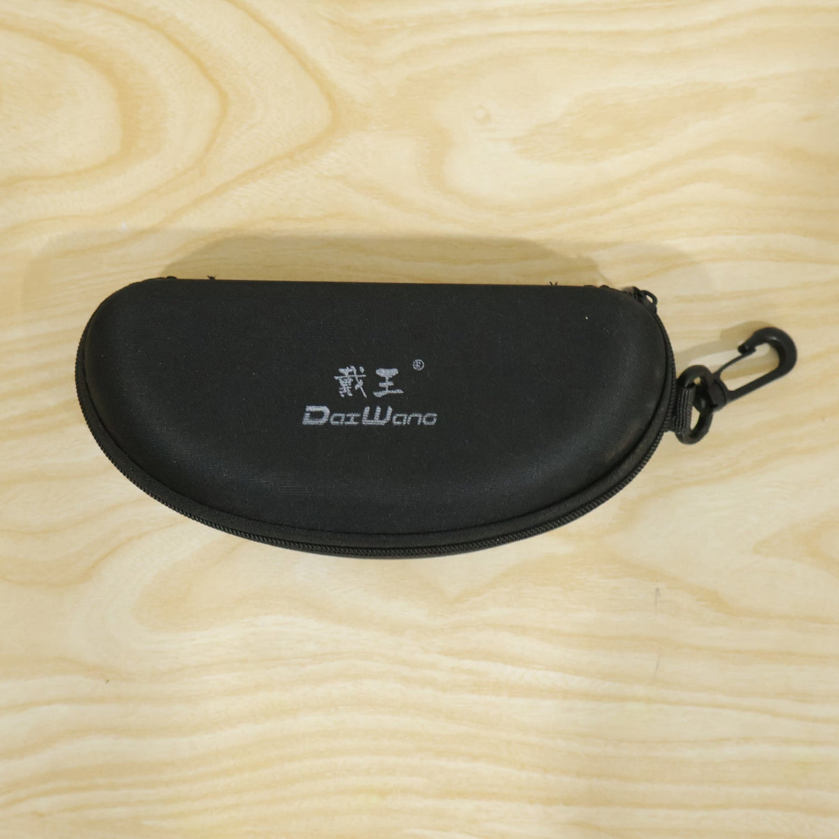 Closed black hard shell sunglasses case with a white logo and clip, on a light wooden surface.