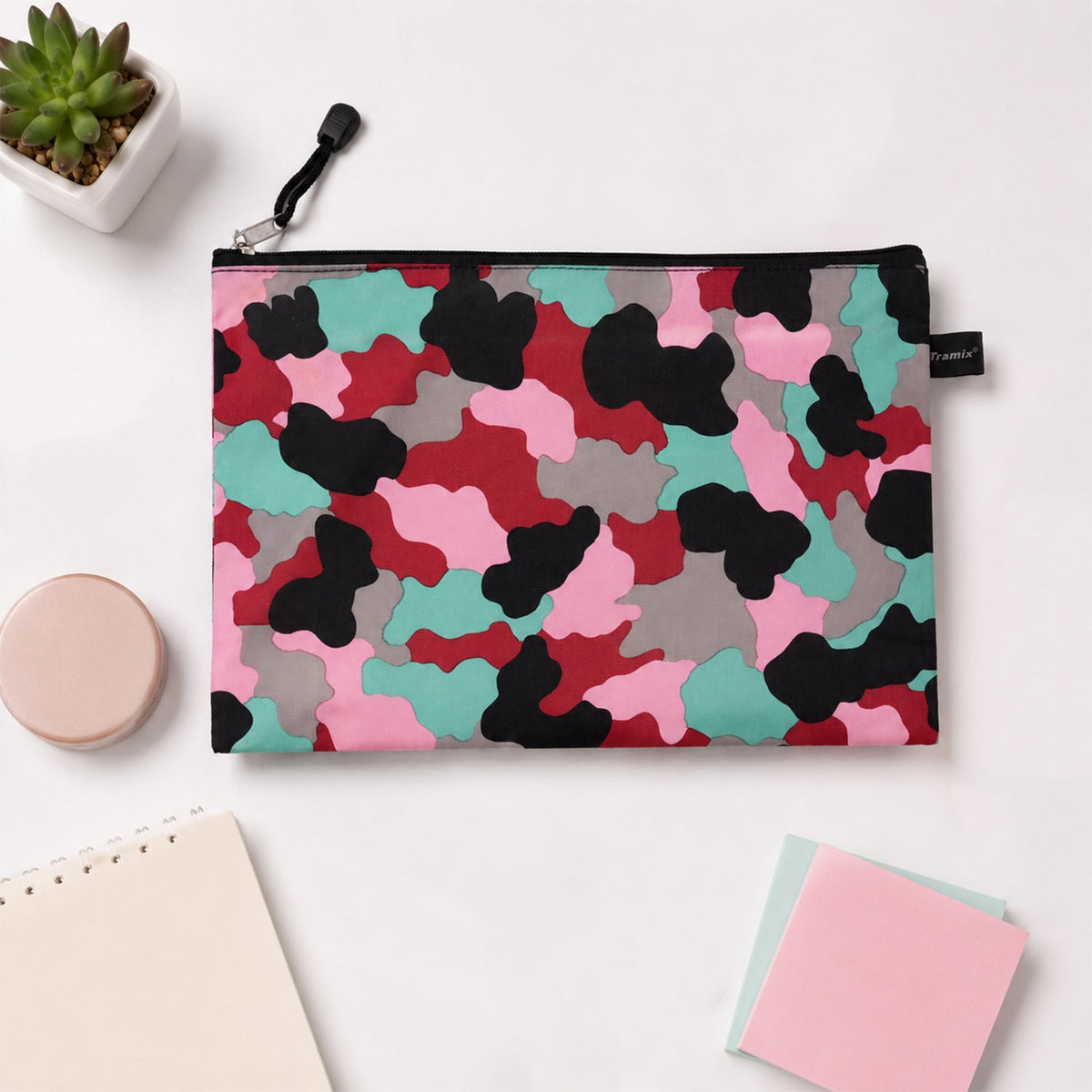 A flat, closed pouch with a pink, red, black, grey, and teal camouflage pattern, resting on a white desk alongside a