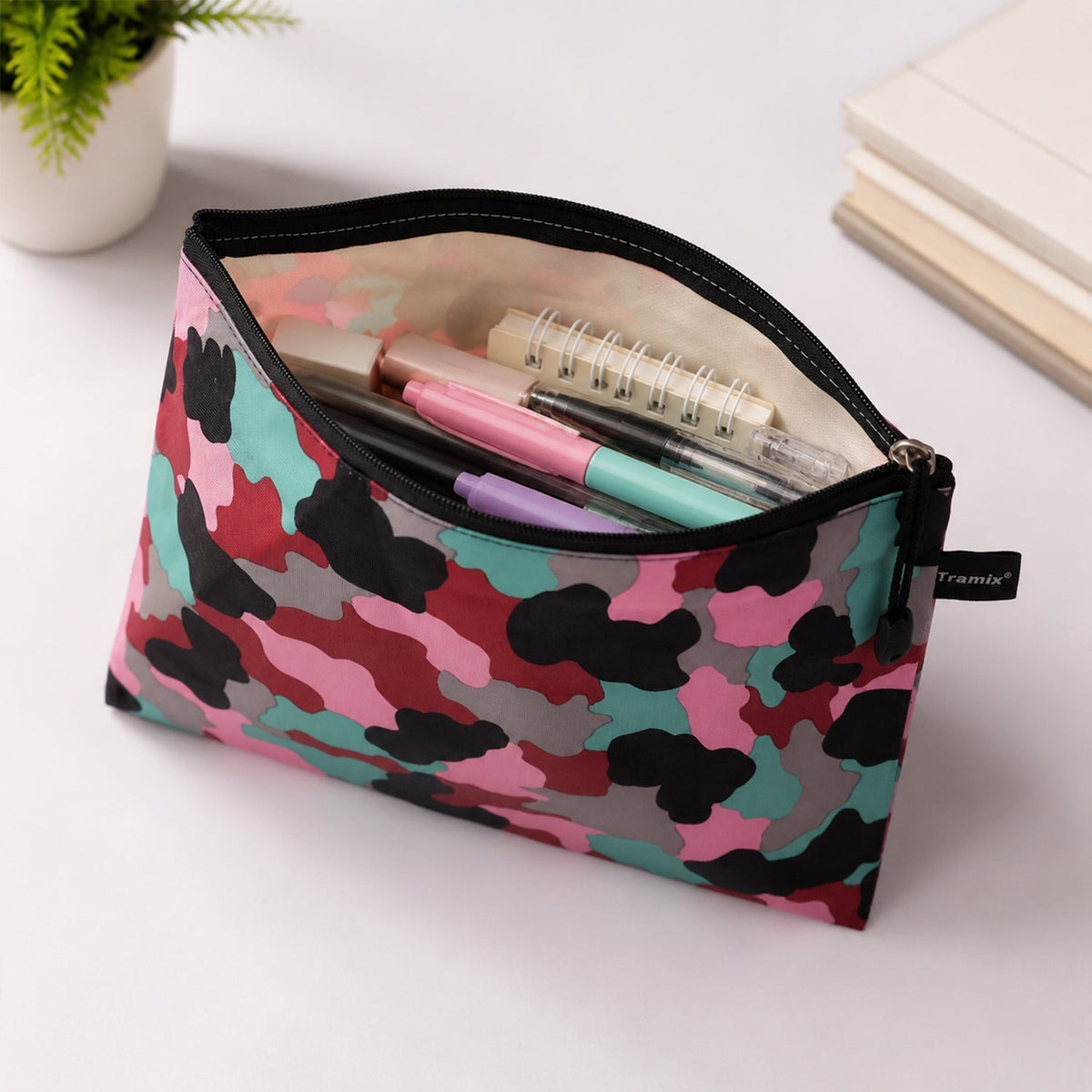 An open pouch with a pink, red, black, grey, and teal camouflage pattern, filled with pens, markers, and a small spiral