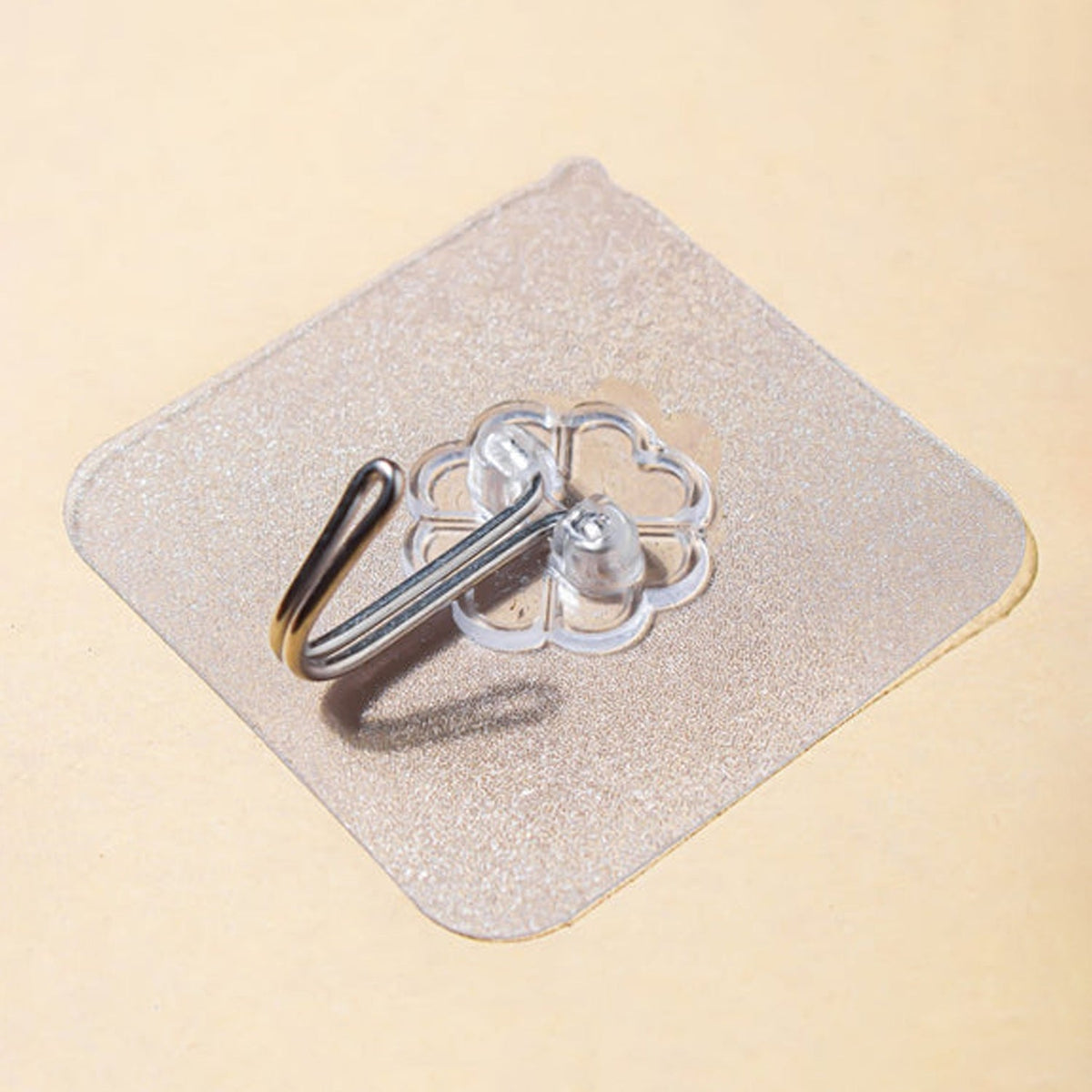 A close-up, angled view of a transparent self-adhesive hook with a silver metal hook on a light brown surface.