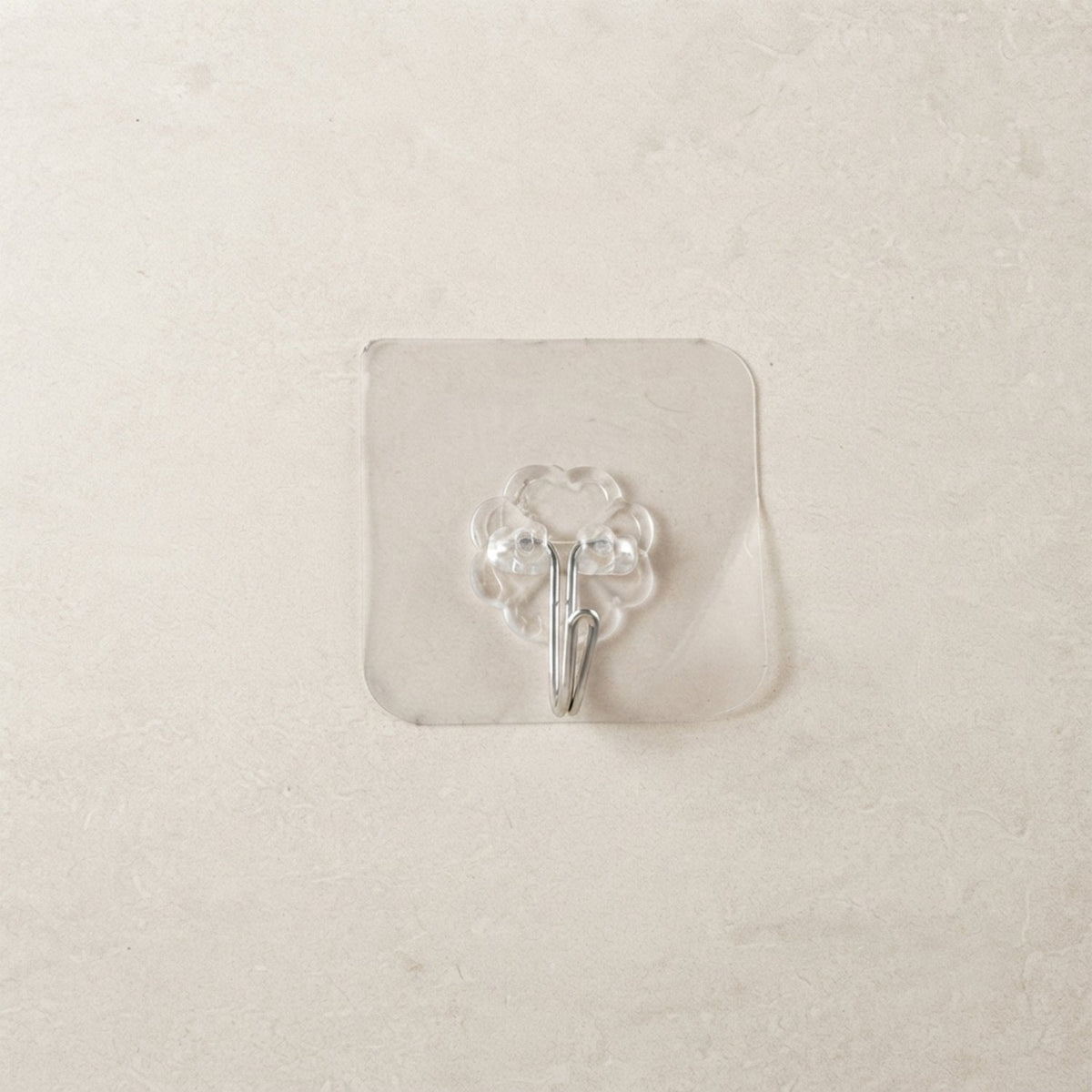 A transparent self-adhesive hook with a silver metal hook centered on a light beige tiled wall.