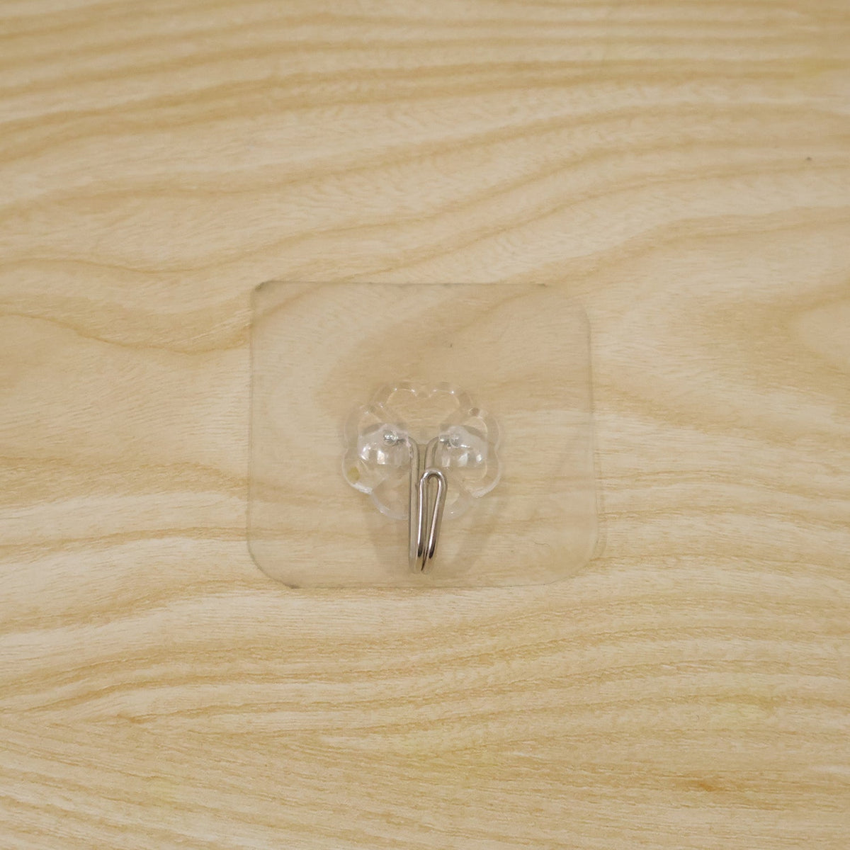 A transparent self-adhesive hook with a silver metal hook centered on a light wooden surface.