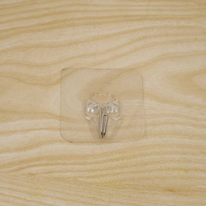 A transparent self-adhesive hook with a silver metal hook centered on a light wooden surface.