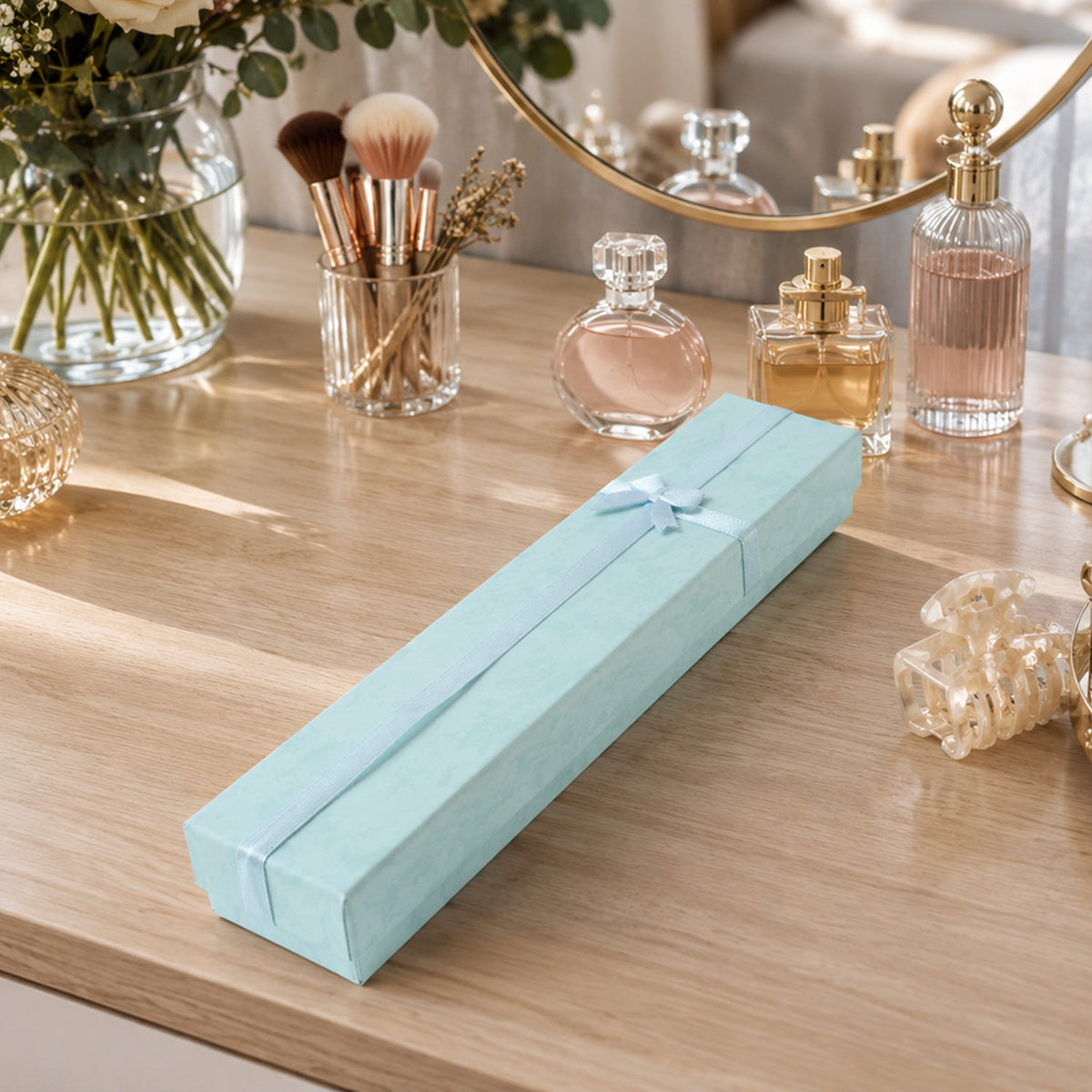 A closed light blue rectangular gift box with a light blue ribbon and bow, on a wooden vanity with perfumes and makeup