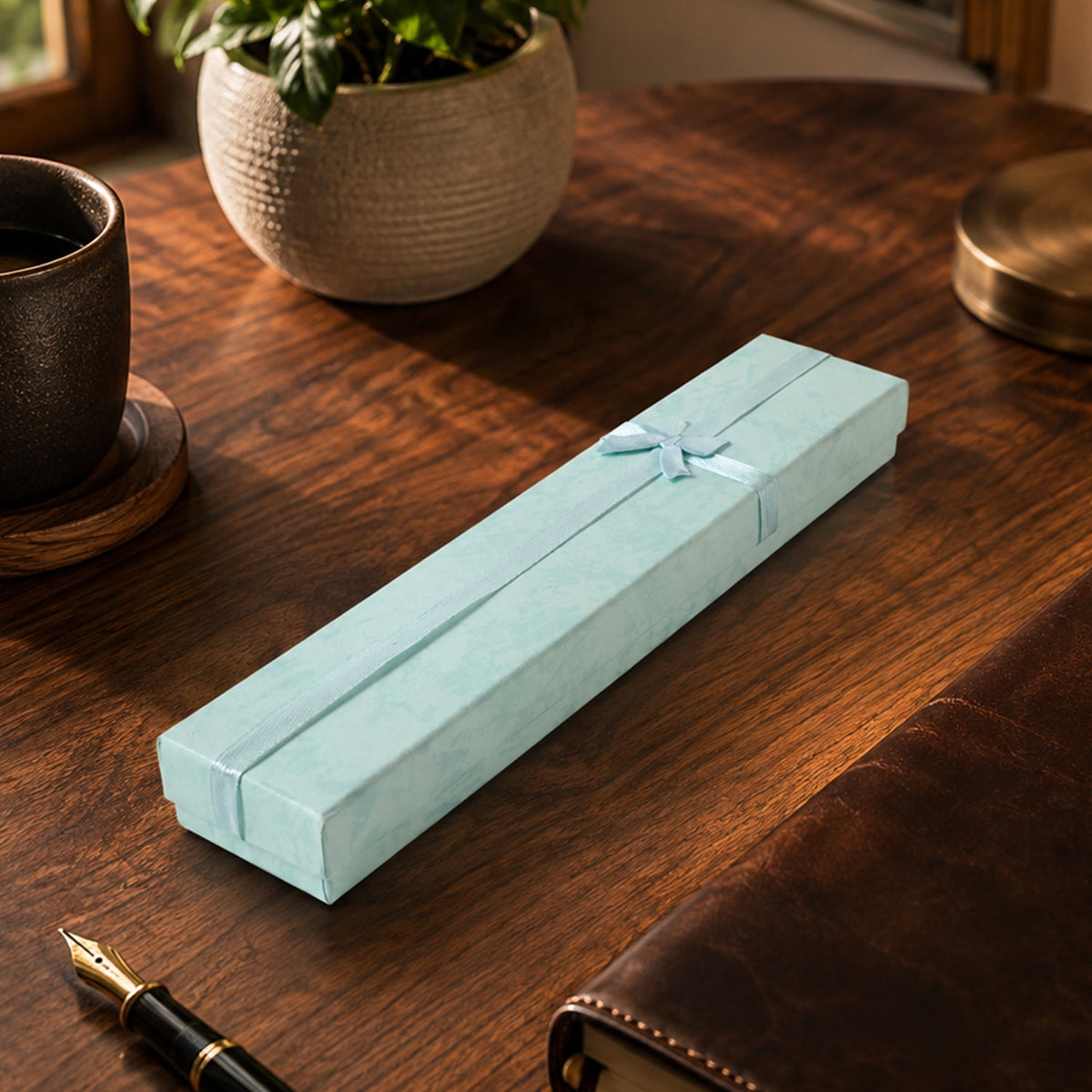 A closed light blue rectangular gift box with a light blue ribbon and bow, on a wooden desk with a coffee cup and a journal.