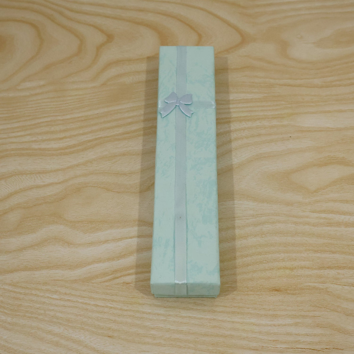 A closed light blue rectangular gift box with a light blue ribbon and bow, on a light wooden surface.