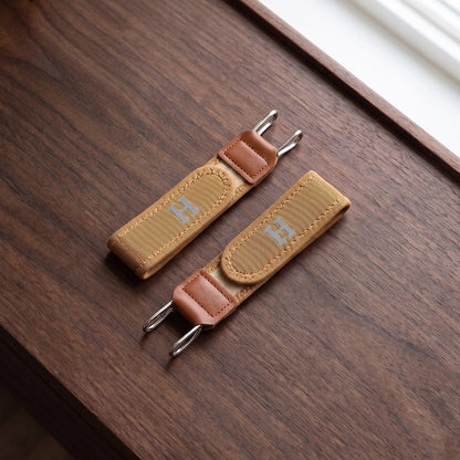 Two tan elastic belt hooks with brown faux leather accents and silver-colored metal hooks, resting on a dark wooden surface.