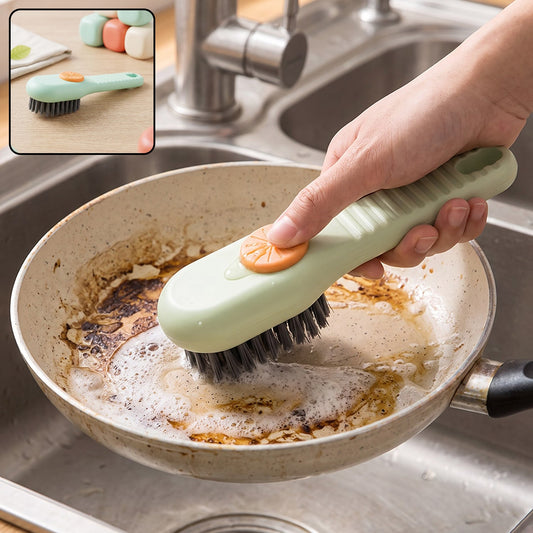 A hand holds a green and orange liquid-dispensing brush, cleaning a dirty frying pan in a stainless steel sink.