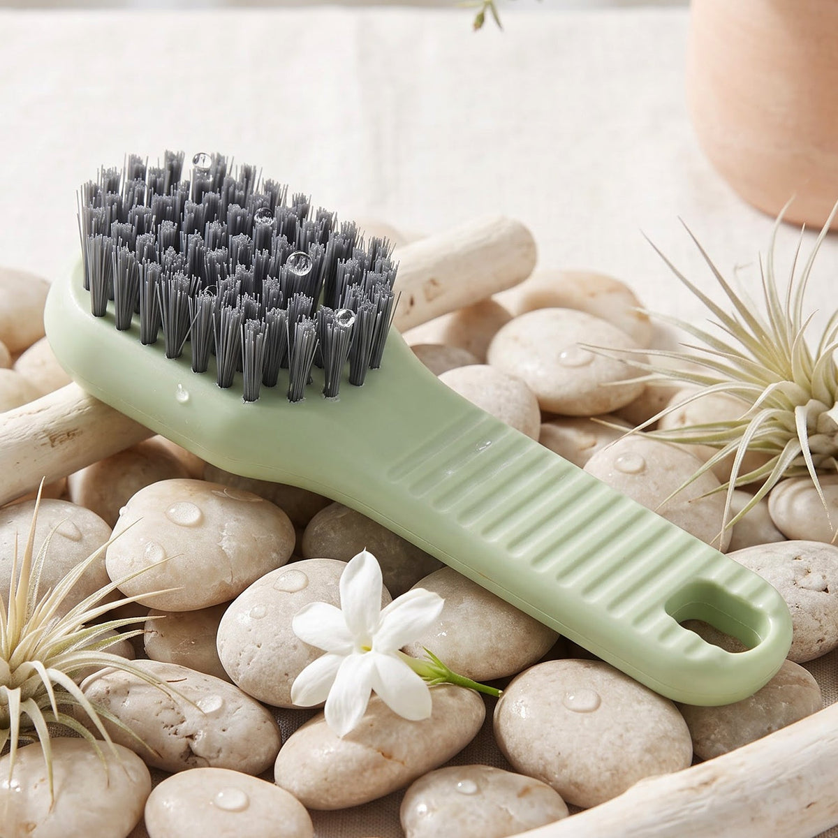 A green and orange liquid-dispensing brush with water droplets on its bristles rests on a bed of light-colored pebbles with