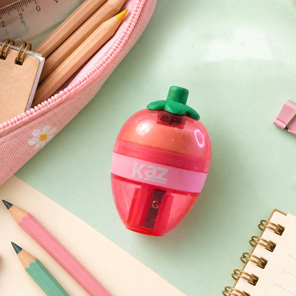 A pink and red strawberry-shaped sharpener with a green stem sits on a desk with pencils and a notebook.