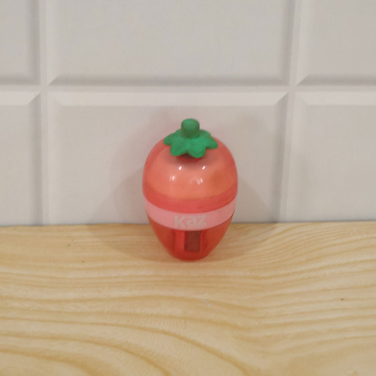 A pink and red strawberry-shaped sharpener with a green stem rests on a light wooden surface against white tiles.