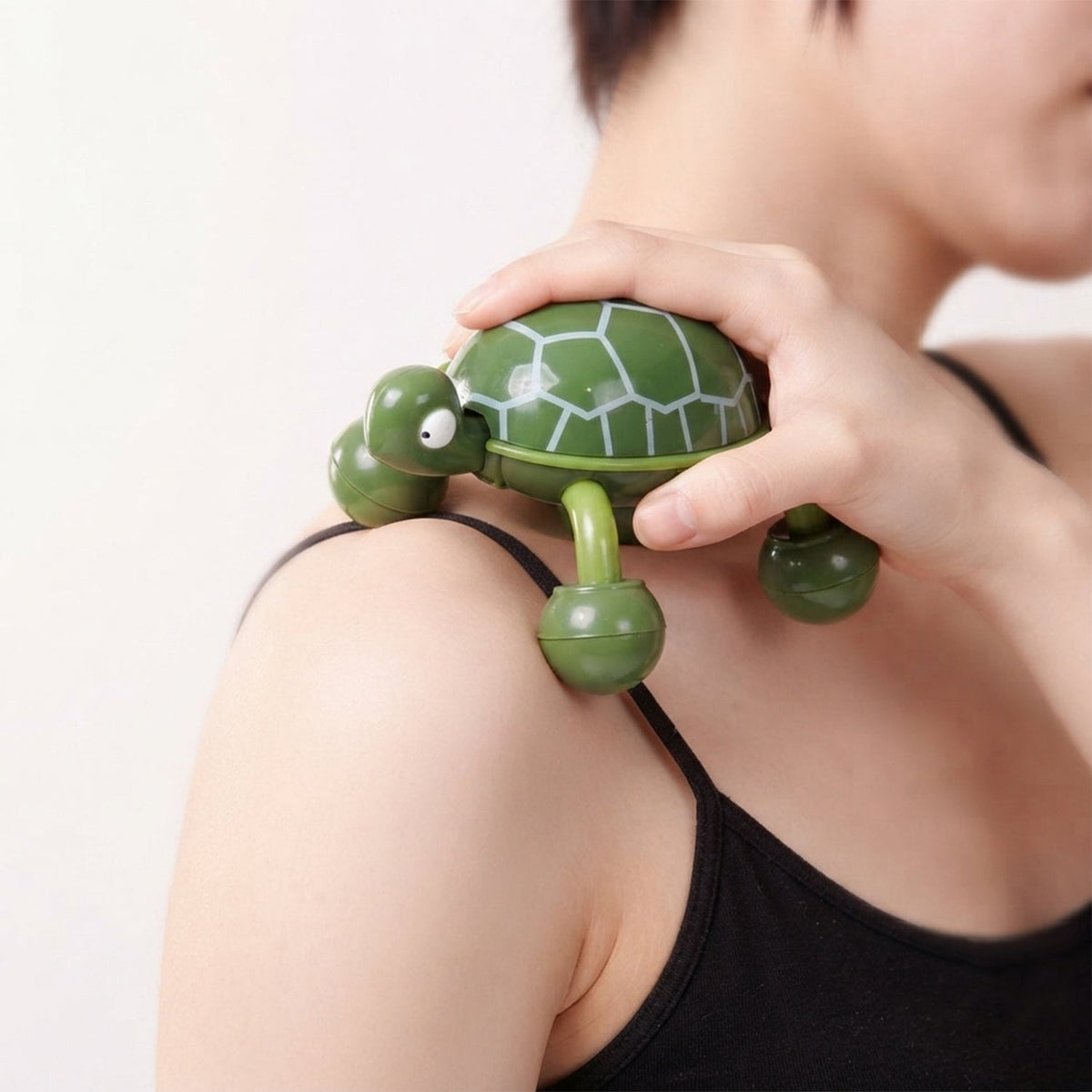 A person's hand manipulates a green turtle massager over their shoulder, highlighting the rolling feet in action.