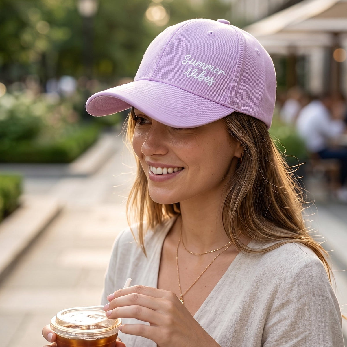 Smiling woman wearing a lavender baseball cap with "Summer Vibes" embroidery, holding a drink outdoors.