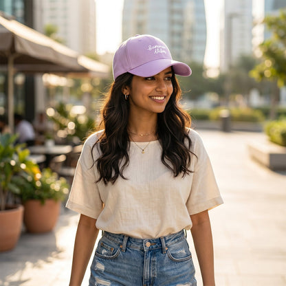 Woman with long dark hair wearing a lavender "Summer Vibes" baseball cap and casual outfit, walking outdoors.