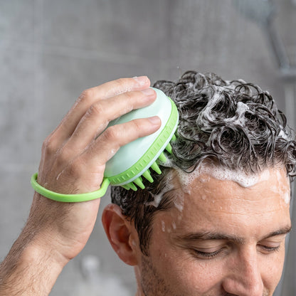 A person's hand is visible, holding a light green silicone scalp massage brush and scrubbing their dark, sudsy hair in a