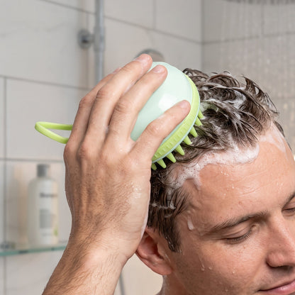 A man massages his wet, dark hair with a light green silicone scalp brush in the shower, with shampoo bottles blurred in the