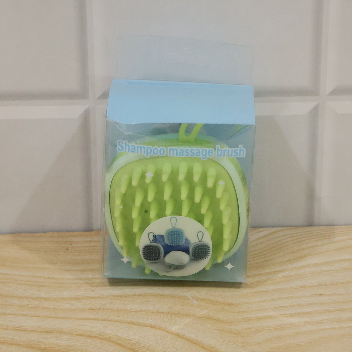 A light green silicone shampoo massage brush is packaged in a clear plastic box with a light blue top, sitting on a wooden