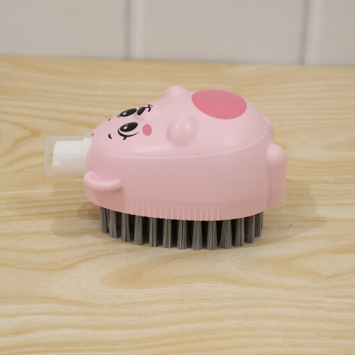 A pink bear-shaped soap dispensing brush with grey bristles rests on a light wooden surface.