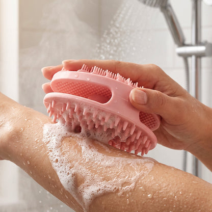 A hand holds a pink silicone body scrubber, scrubbing a soapy arm under a shower head.