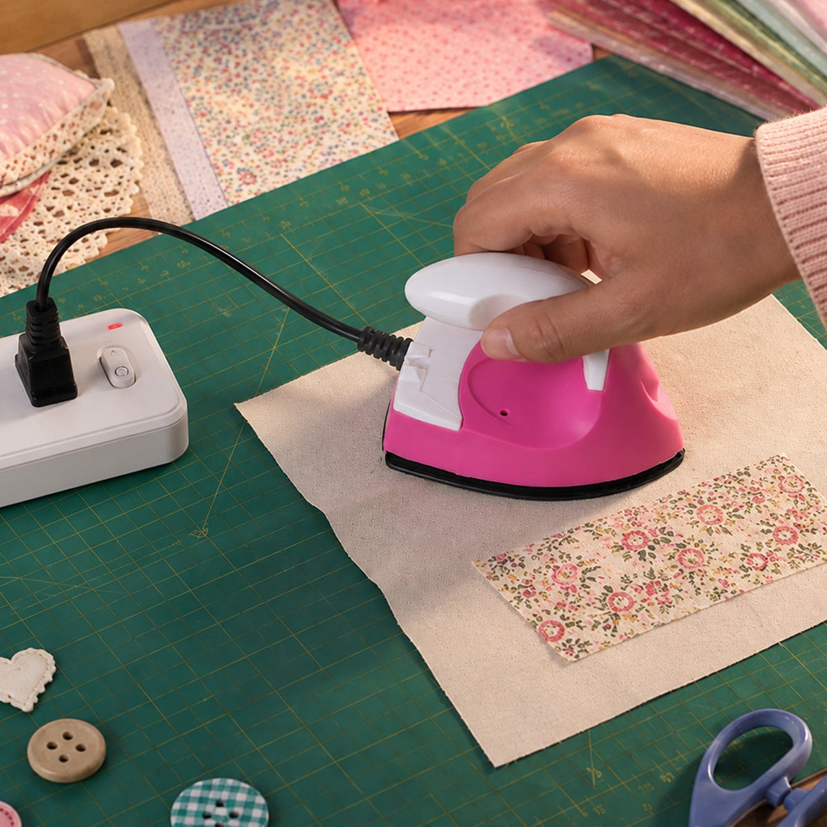 A hand uses a pink and white mini iron on fabric, with the iron plugged into a white power strip.