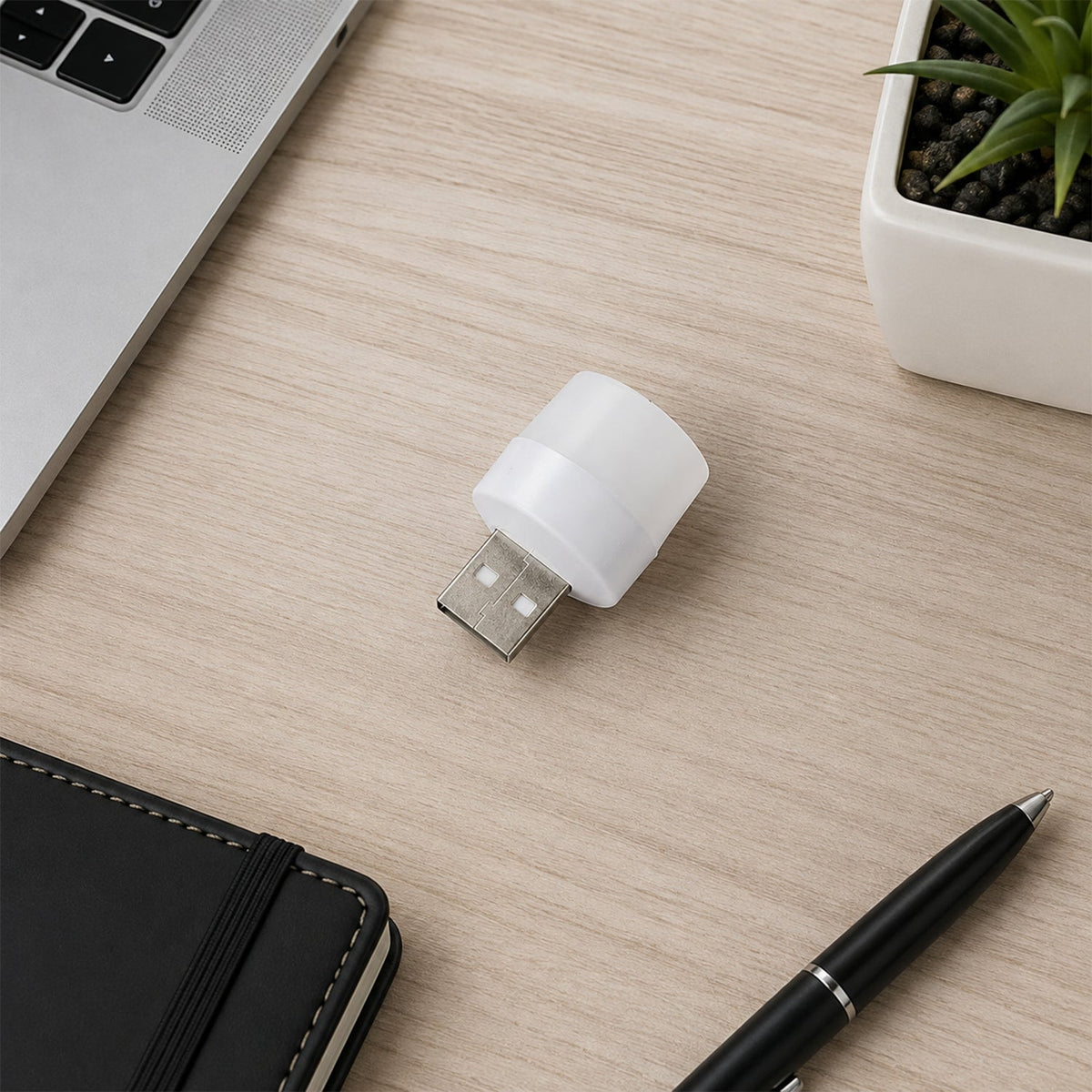 A white USB mini light bulb resting on a light wooden desk next to a laptop, notebook, pen, and small potted plant.