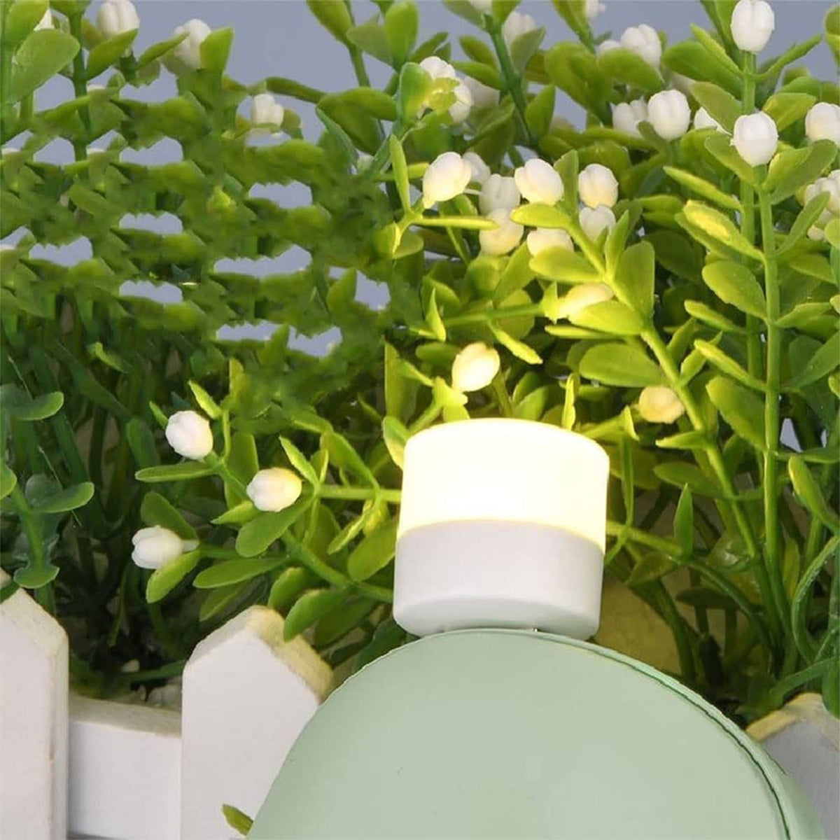 A white USB mini light bulb emitting a warm glow, placed among green faux plants with small white flowers.