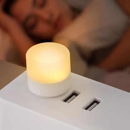 A warm white USB night light plugged into a white power strip, with a sleeping person blurred in the background.