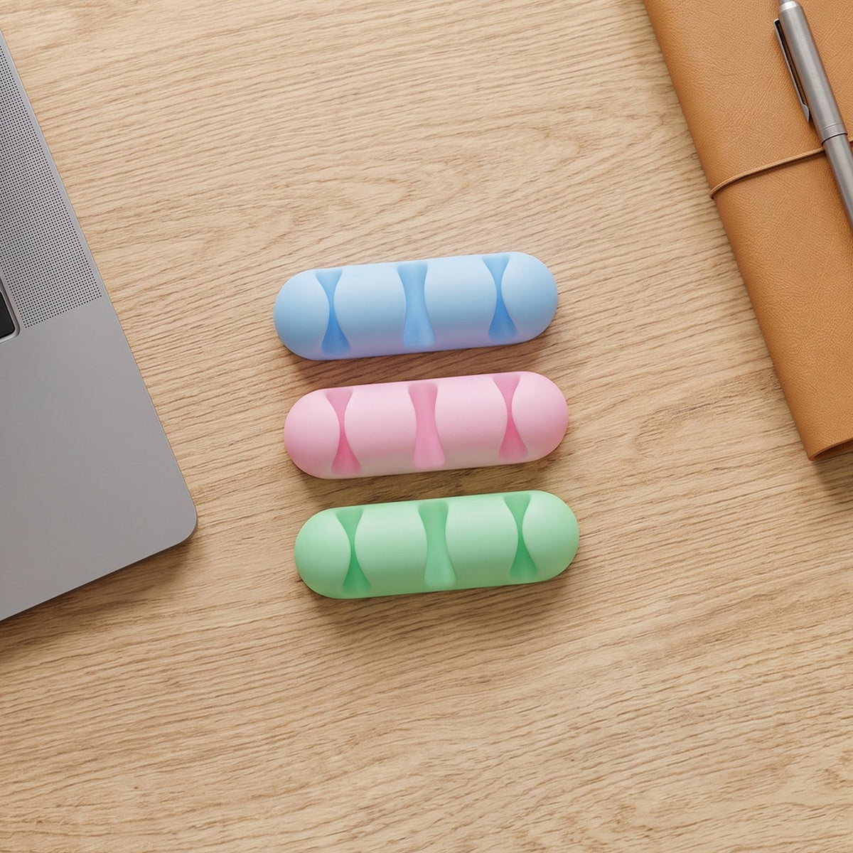 Three silicone cable organizers in blue, pink, and green are arranged on a light wooden desk next to a laptop and notebook.
