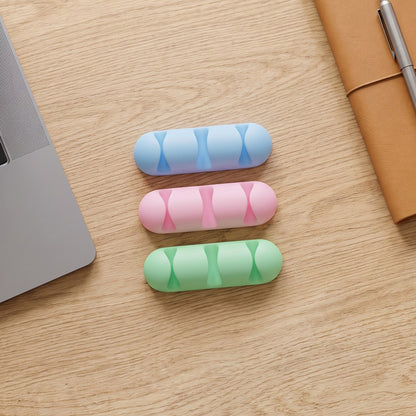 Three silicone cable organizers in blue, pink, and green are arranged on a light wooden desk next to a laptop and notebook.