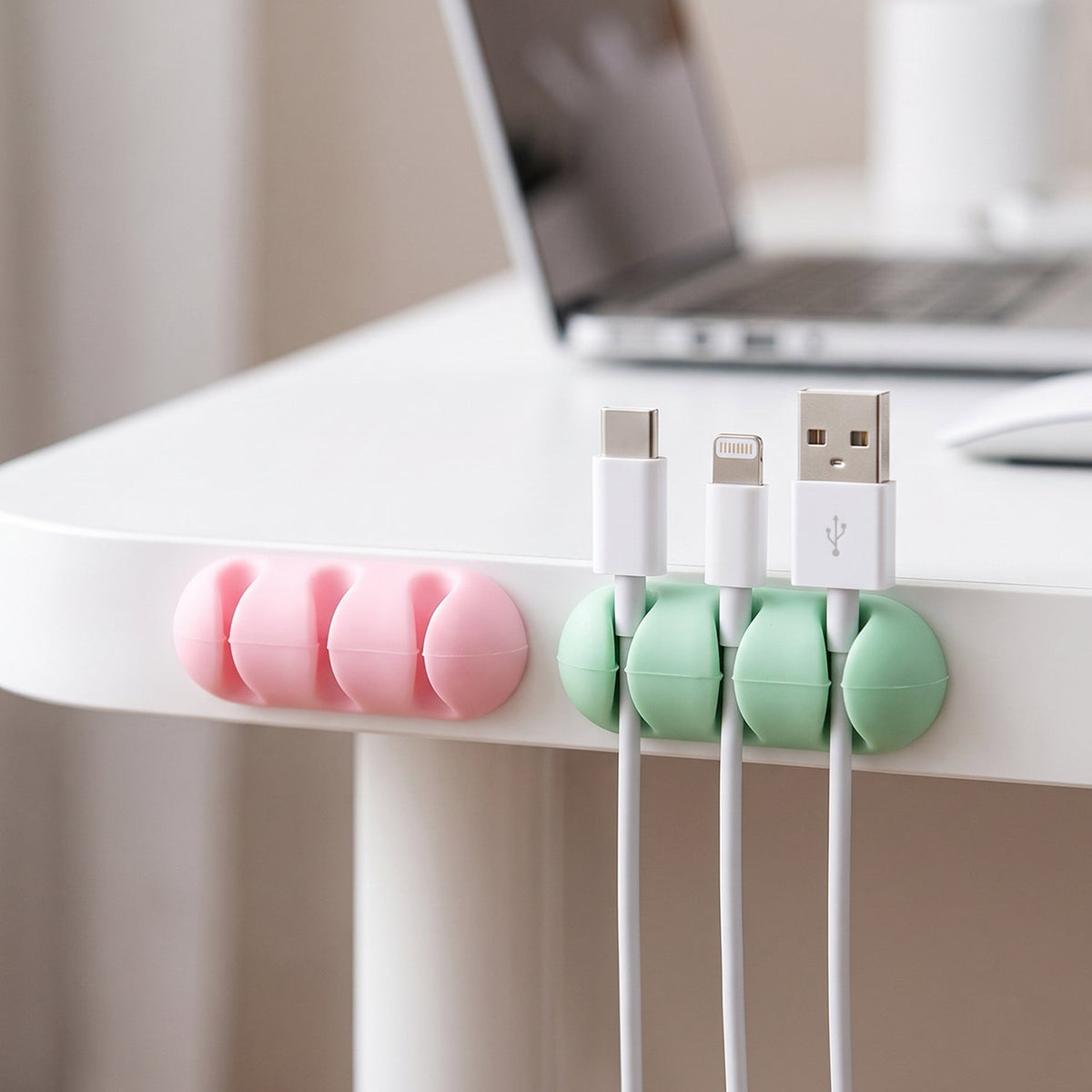 A pink and a green silicone cable organizer are attached to a white desk, holding three charging cables, with a laptop in