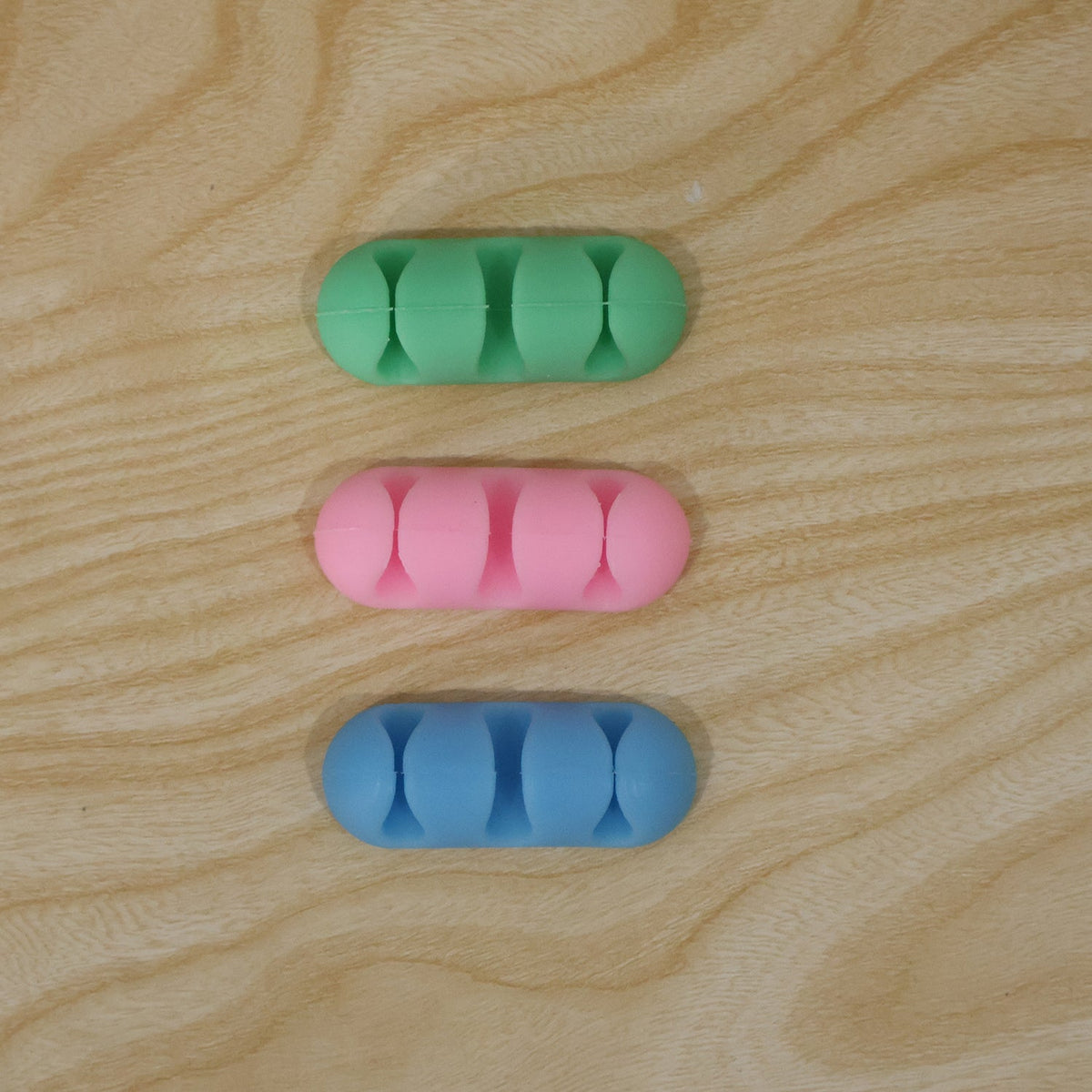 Three silicone cable organizers in green, pink, and blue are arranged vertically on a light wooden surface.