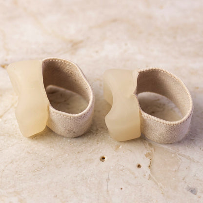 Two beige elastic toe separators with clear gel pads, viewed from the side, sitting on a light-colored stone surface.