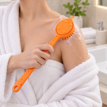 A person in a white robe uses an orange silicone back scrubber with soap suds on their shoulder in a bathroom.