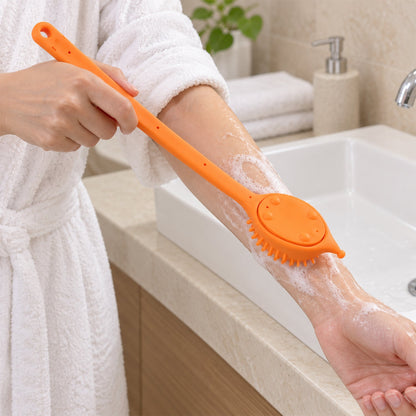 A person in a white robe uses an orange silicone back scrubber on their arm, creating soap suds, in a bathroom.