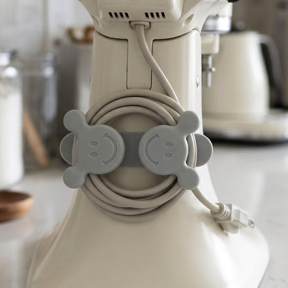 A gray silicone cord organizer with two smiling faces, holding a coiled light gray appliance cord on a white stand mixer.