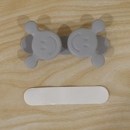 A gray silicone cord organizer with two smiling faces and a white adhesive strip on a light wood surface.